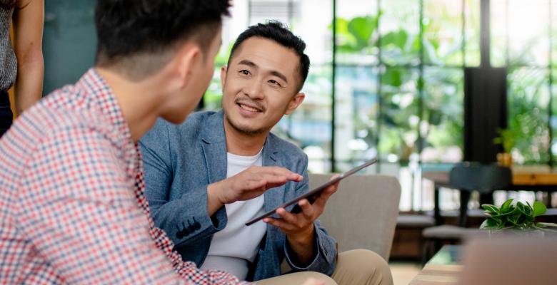 Two men sitting and working together as one explains something to the other, as he holds a digital tablet.